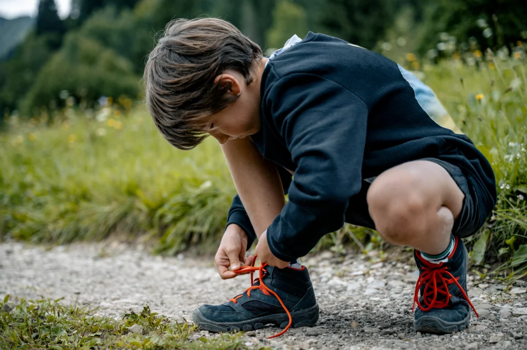 Young boy outdoors crouching on gravel path tying red shoelaces on dark hiking shoes.