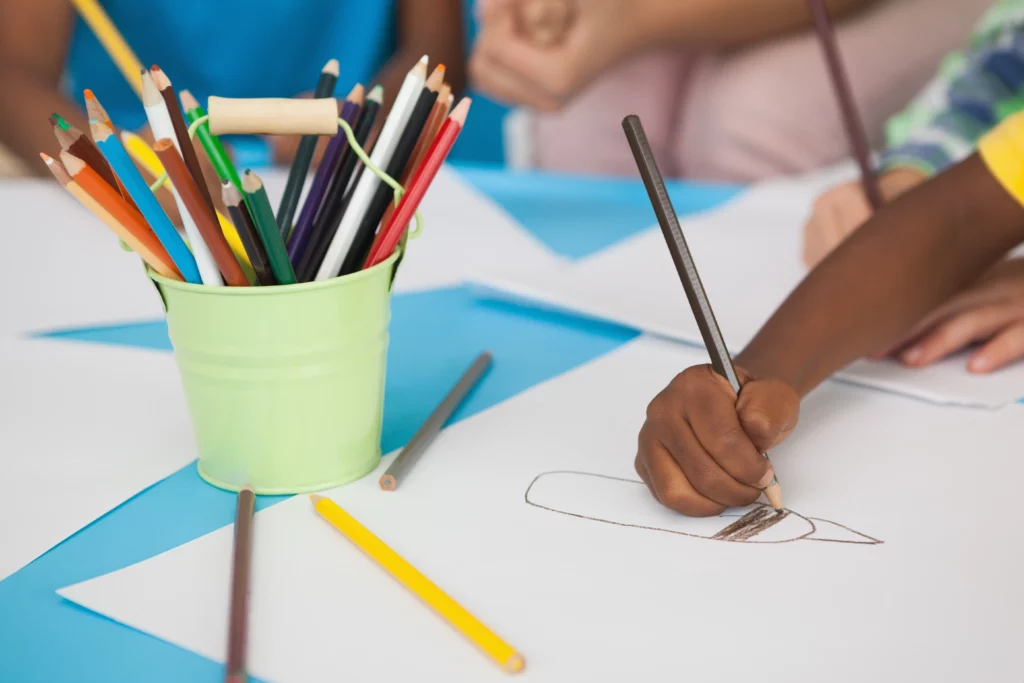 Child drawing with brown colored pencil on white paper, green container with pencils nearby.