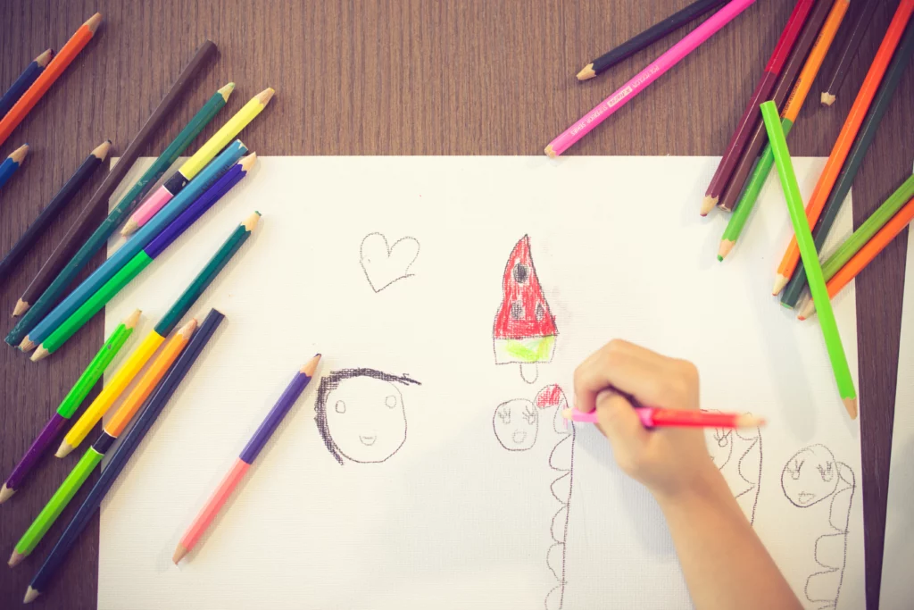 Child drawing faces, a heart, and a red-green object on white paper with scattered colored pencils.