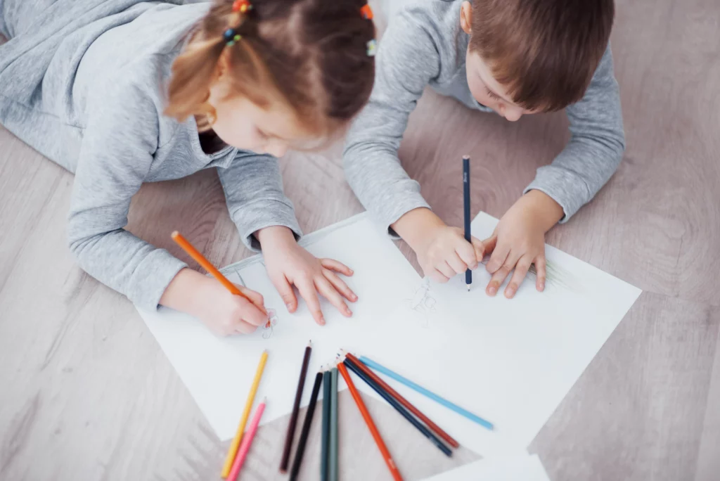 Two children lie on the floor drawing with colored pencils on white paper sheets.
