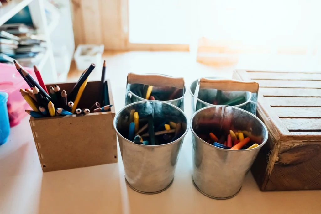 Wooden container and three metal buckets with colored pencils and crayons on white surface.
