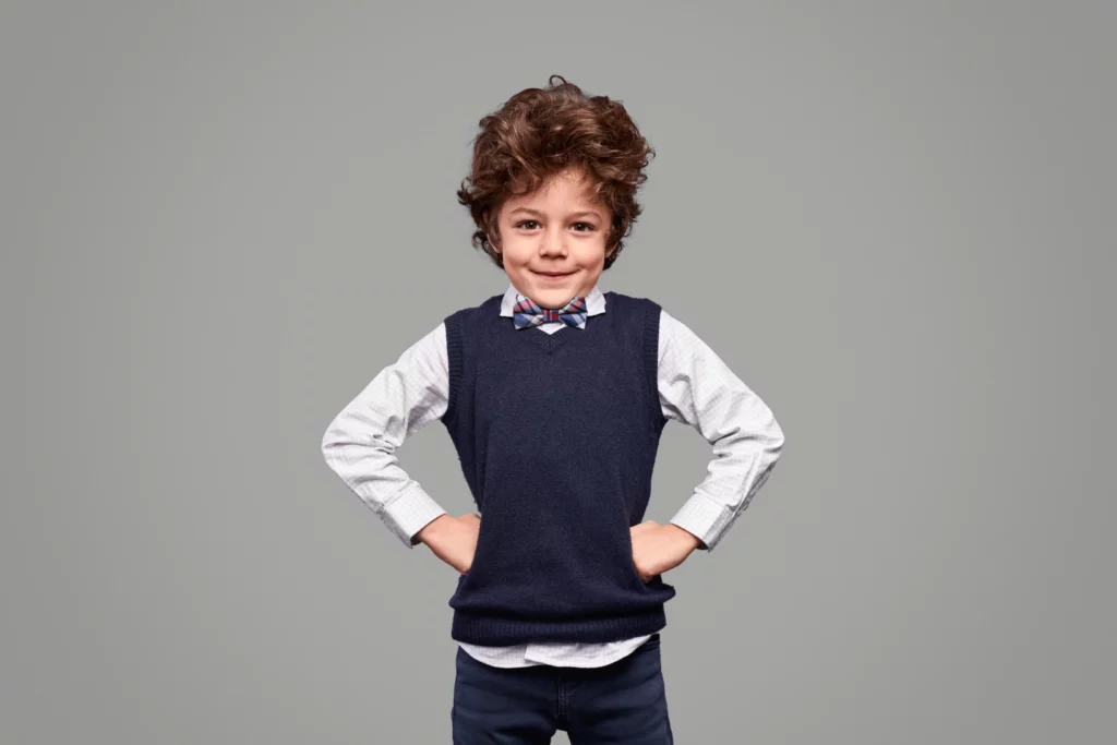 Young boy with curly hair wearing a white checkered shirt, navy sweater vest, and plaid bow tie.