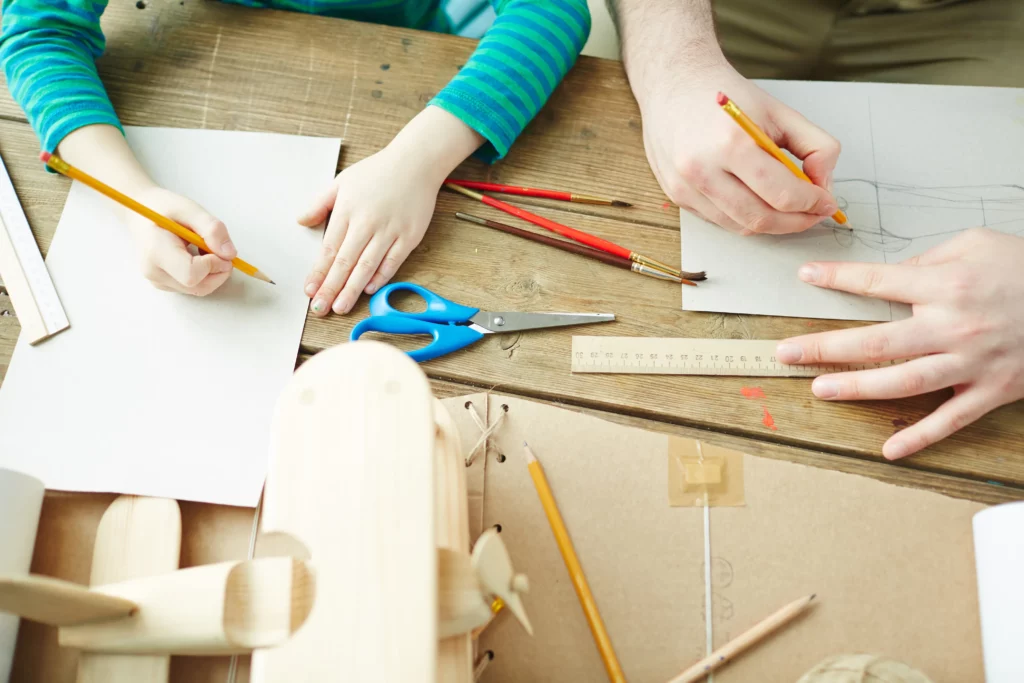 Two people sketching on paper at a table with art supplies and a wooden model airplane.