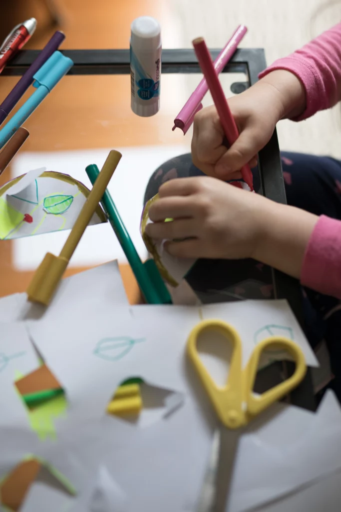 Child coloring cut-out paper shapes on glass table with markers, glue stick, and yellow scissors.