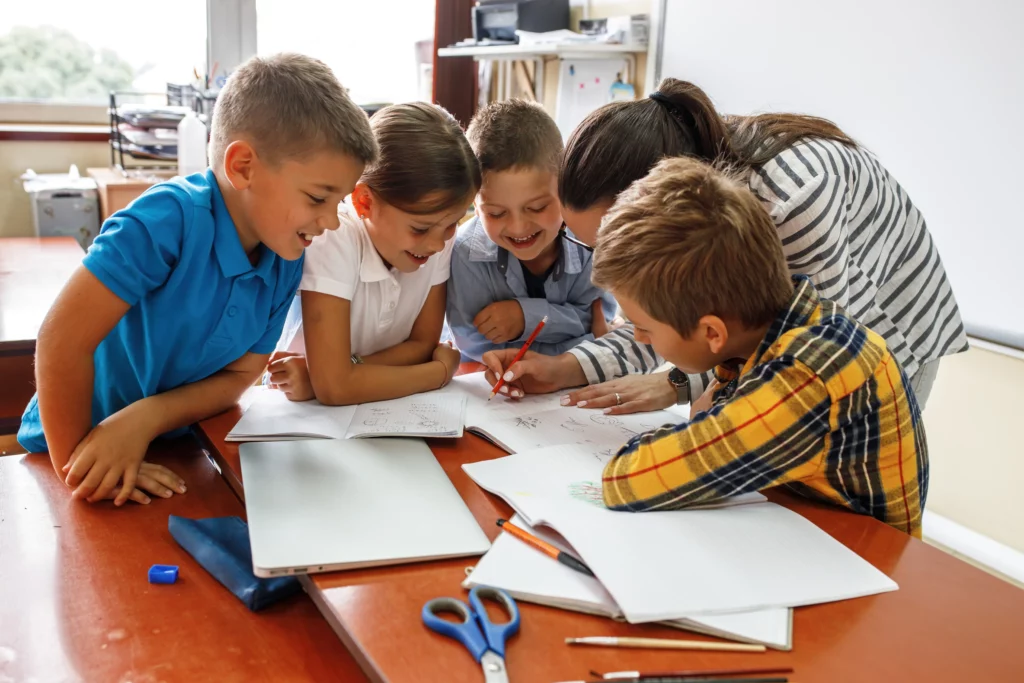 Teacher and four children gathered around a table with notebooks, pencils, scissors, and a closed laptop.