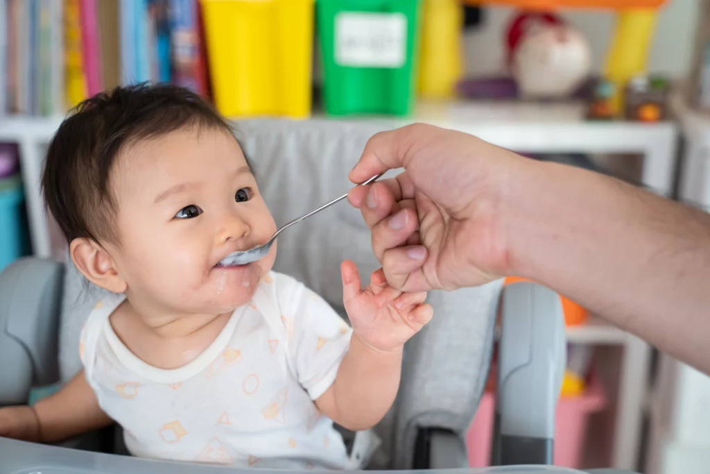 Baby in high chair being fed with a spoon by an adult, food around mouth.