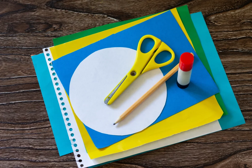 Stack of colorful paper with yellow scissors, pencil, glue stick, and white circle cutout on wood.