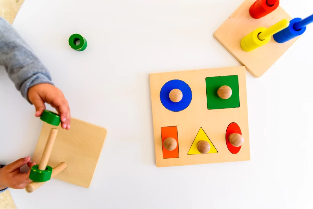 Child assembling wooden stacking toy with green rings near shape sorter and colorful stacking rings on table