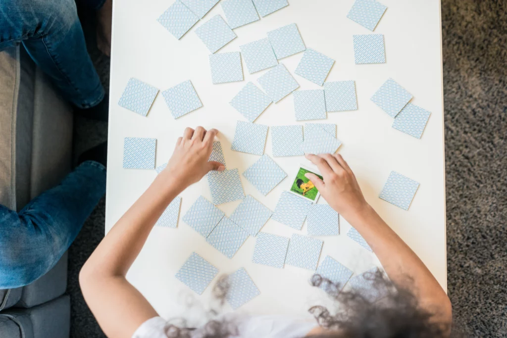 Two people playing a memory matching card game on a white table with scattered cards.