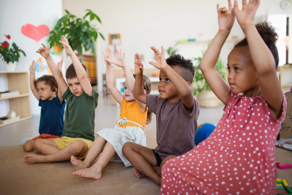 Young children sitting cross-legged on the floor indoors with their hands raised.