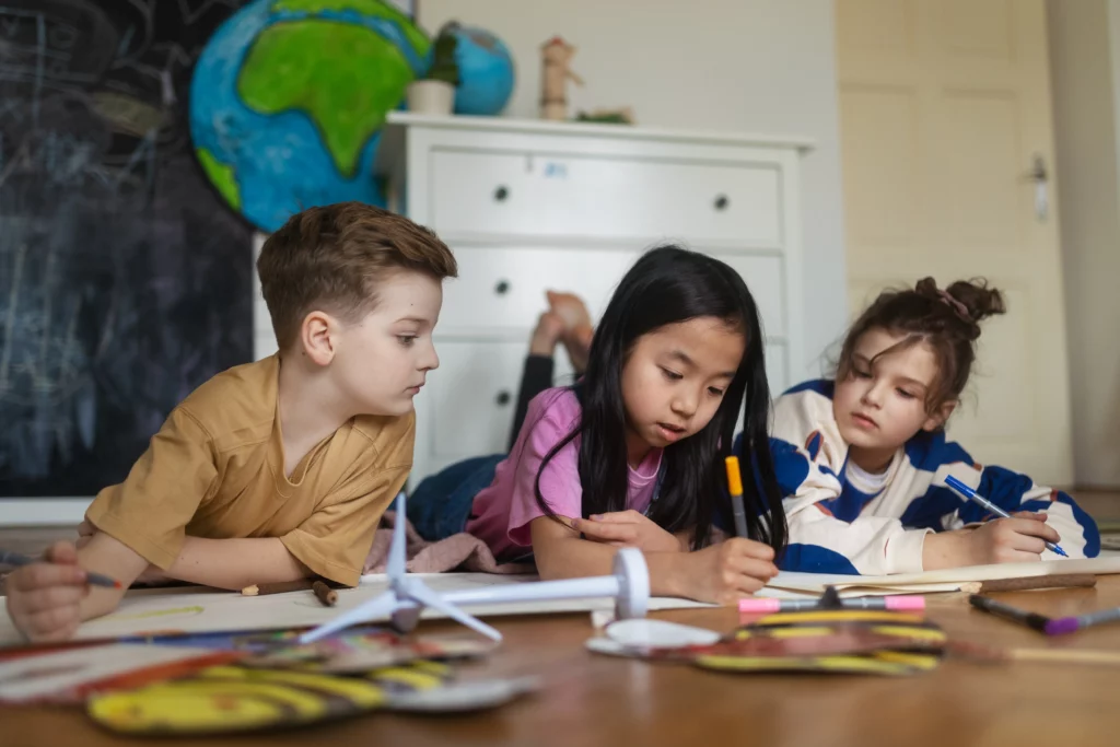 Three children lying on the floor drawing with art supplies near a model wind turbine and globe.