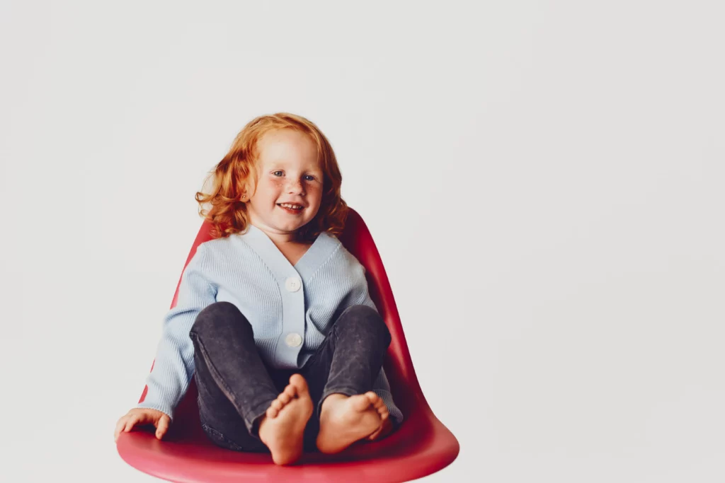 Young red-haired child with freckles sitting barefoot in red chair wearing blue cardigan and dark pants.