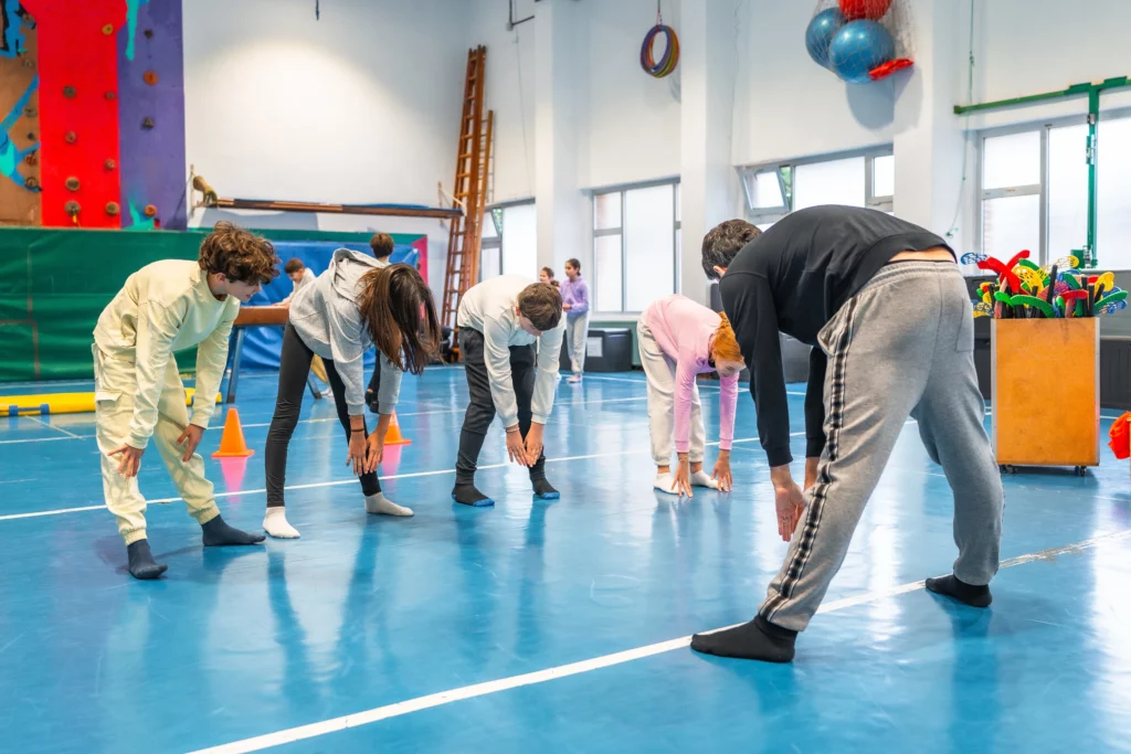 Children in gym stretching forward to touch their toes during a guided exercise session.