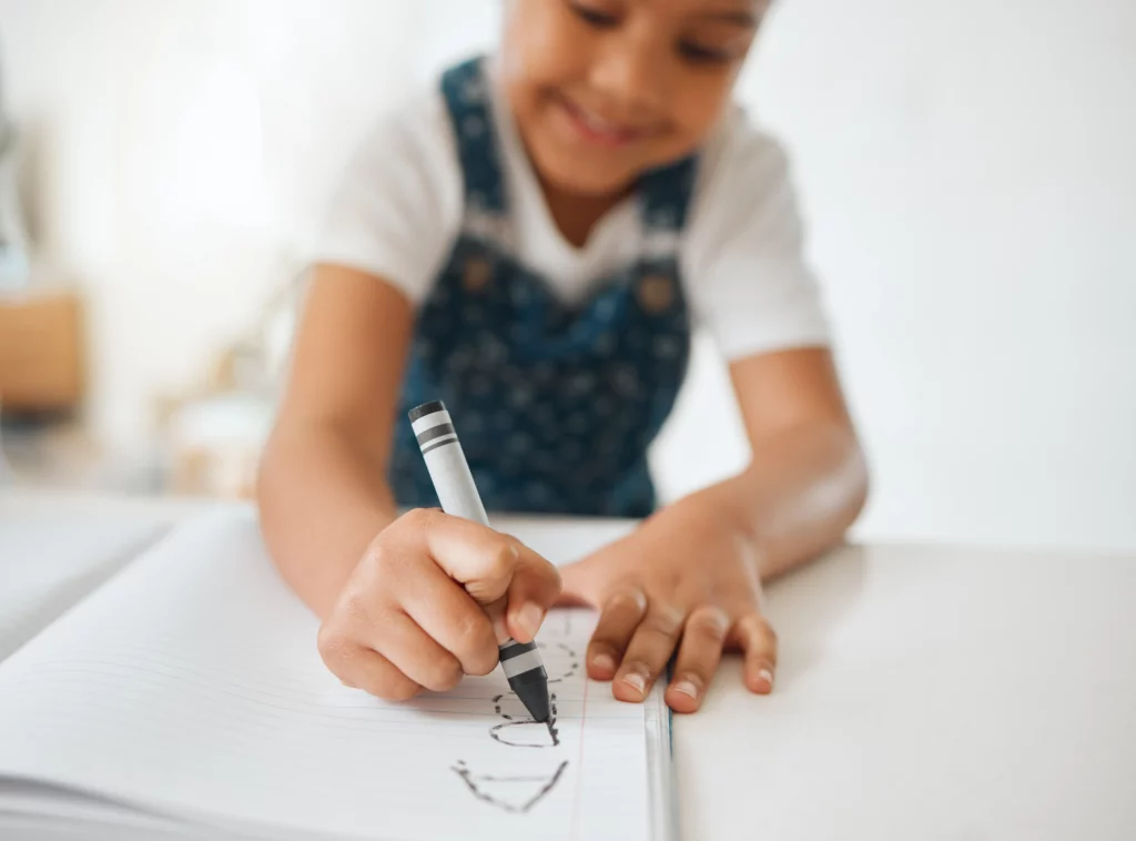 Child drawing with a black crayon on a lined notebook wearing a white shirt and floral denim overalls
