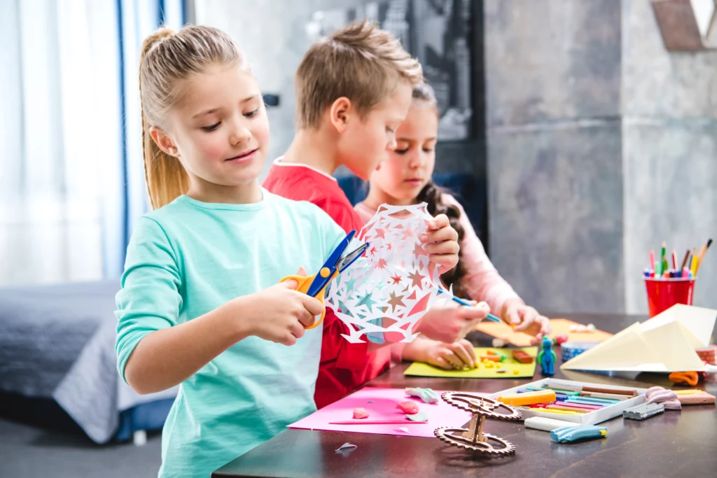 Three children engaged in arts and crafts, including paper cutting and drawing, at a table.