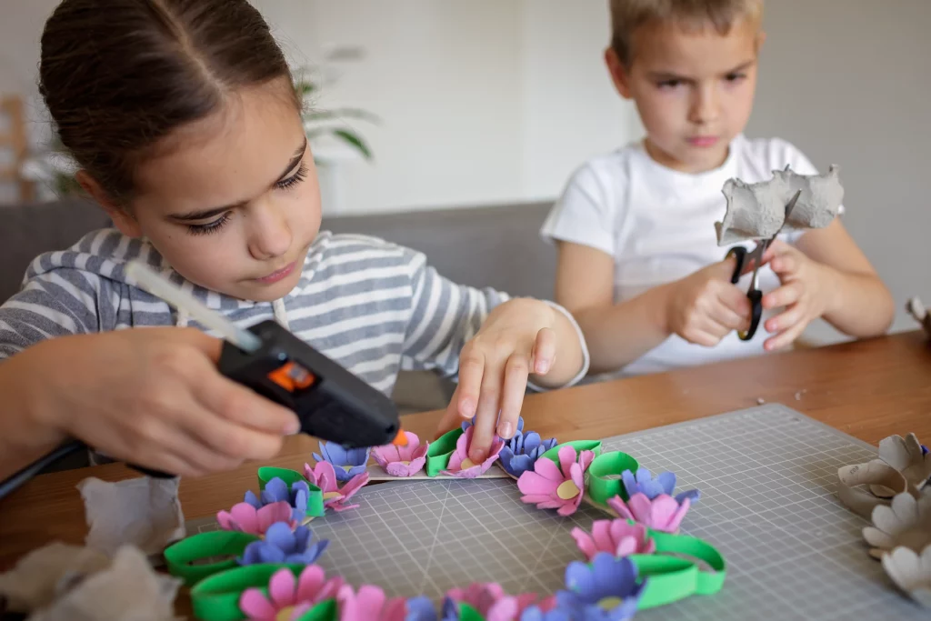 Two children crafting with hot glue and scissors using colorful paper flowers and egg cartons.