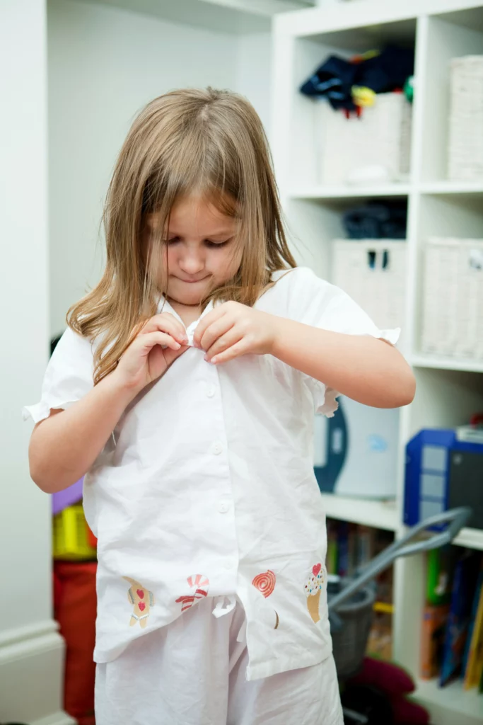Young girl buttoning a white shirt with candy and ice cream illustrations indoors.