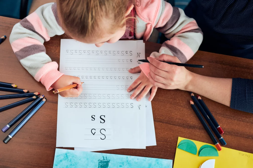 Child practicing writing the letter S on a worksheet with adult guidance at a wooden table.