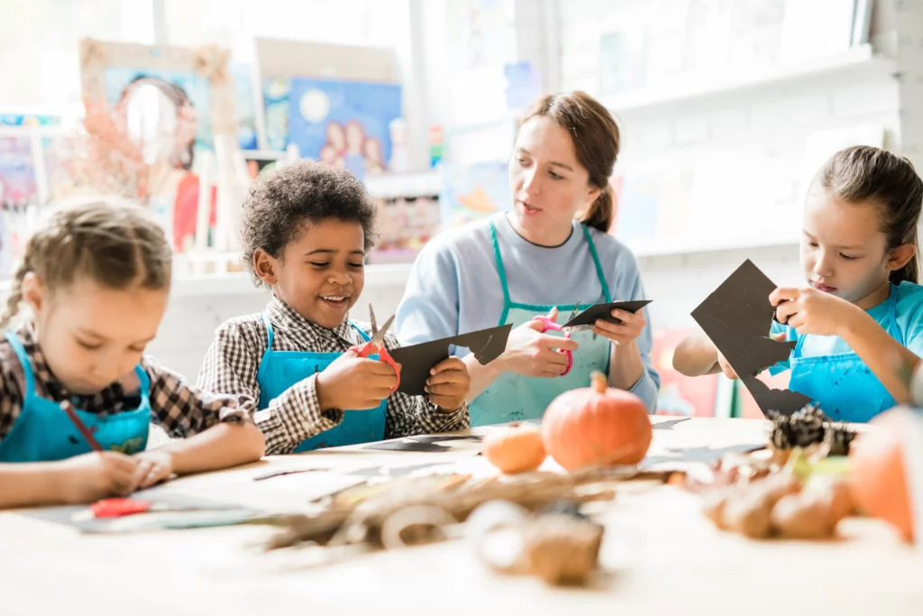 Children and teacher cutting black paper for crafts at a table decorated with pumpkins and pinecones.