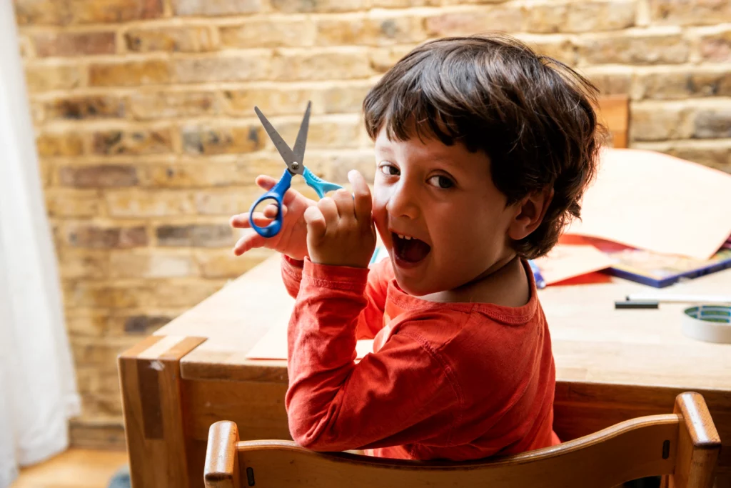 Child in a red shirt sitting at a wooden table holding blue scissors with a brick wall background.