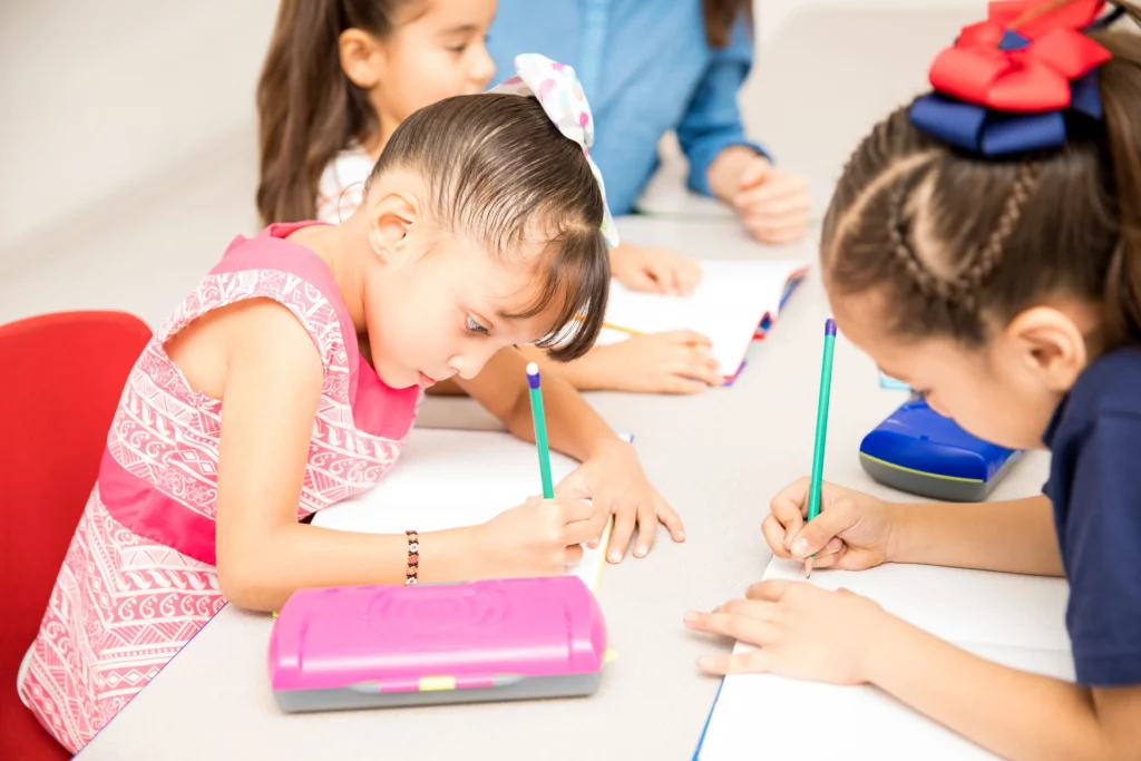 Three young girls focused on writing in notebooks with pencils at a table with stationery items.