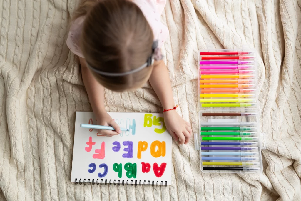 Child lying on a blanket coloring colorful alphabet letters on a spiral sketchpad with markers nearby