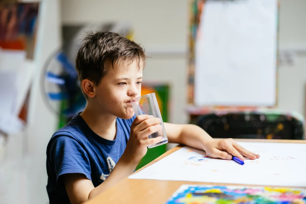 Boy drinking water at a table with drawings, holding a blue pen in one hand.