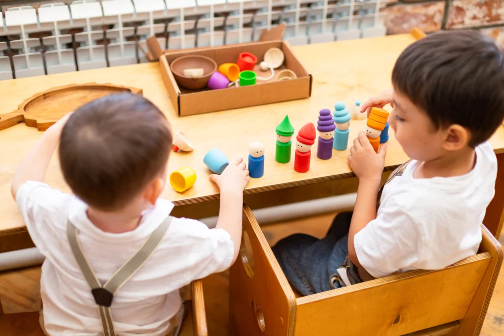 Two children sitting at a wooden table playing with colorful wooden stacking toys and a box of pieces.