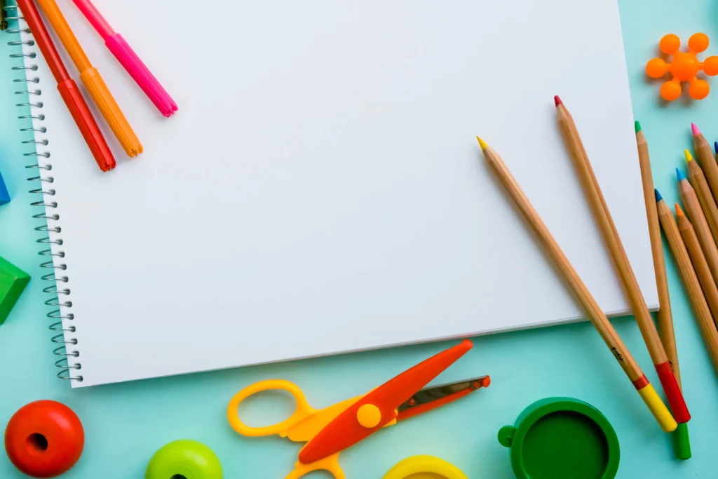 Blank spiral sketchbook with colored pencils, markers, scissors, and paint containers on blue surface