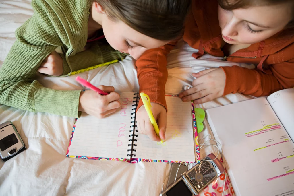 Two girls lying on a bed writing and highlighting in a notebook with a textbook and flip phone nearby.