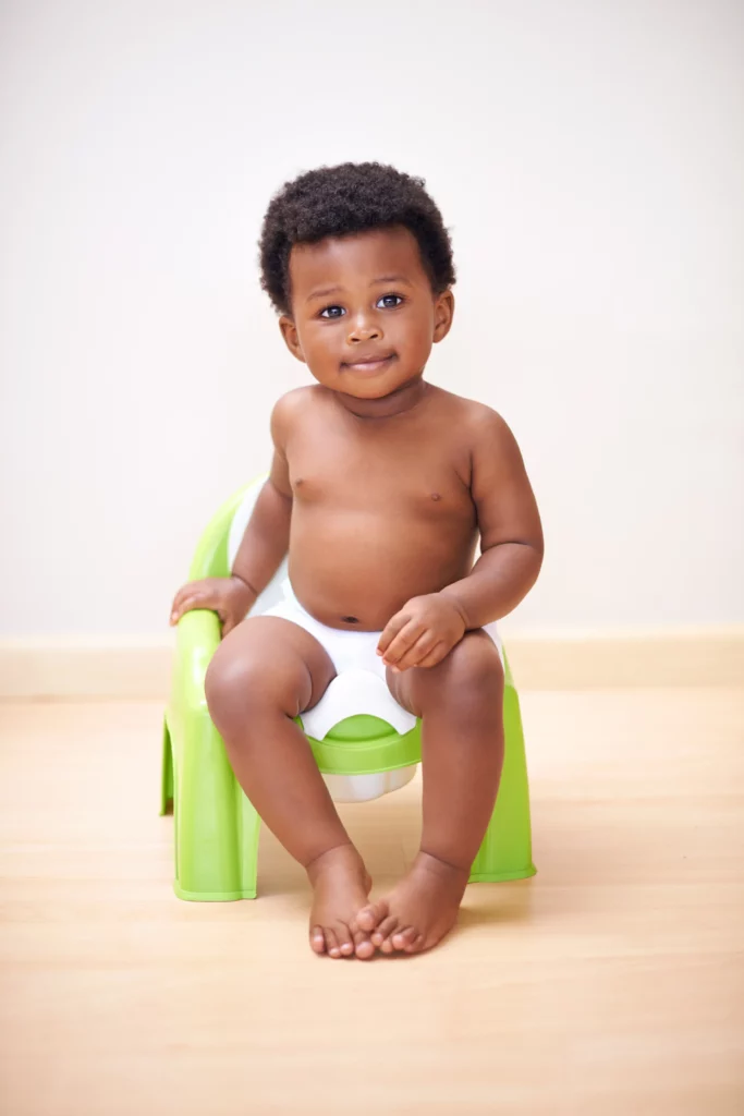 Toddler in a diaper sitting on a green potty chair on a wooden floor with a plain background