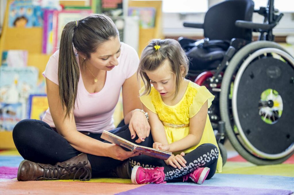 Woman and young girl sitting on carpet reading a book, wheelchair in the background.