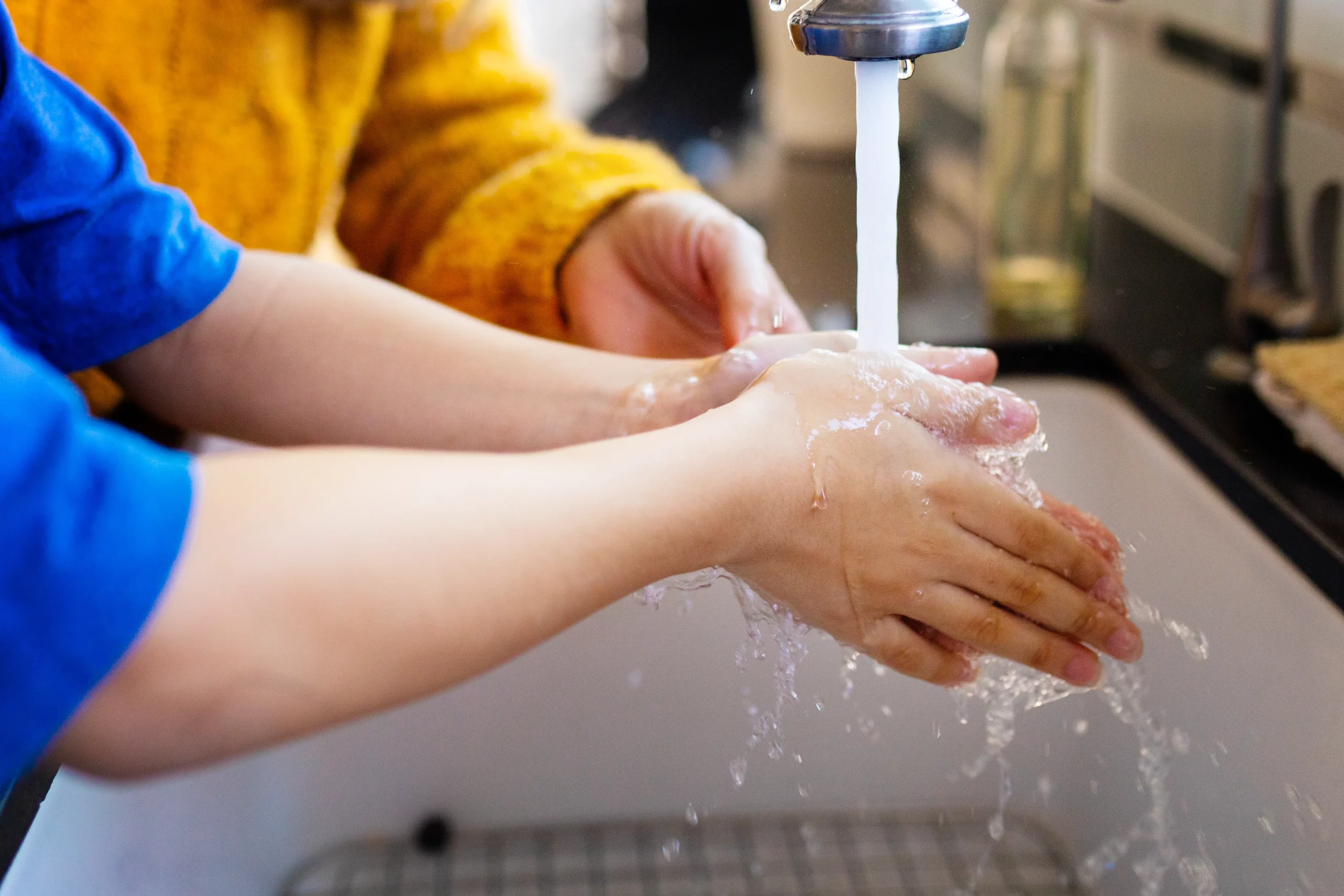 Hands of a child being washed under running water with adult helping at a kitchen sink