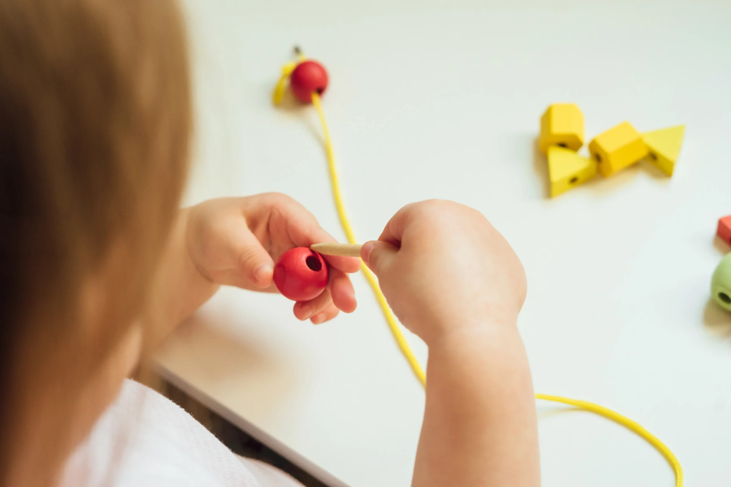 Child threading large red bead with wooden stick on white table with yellow string and beads.