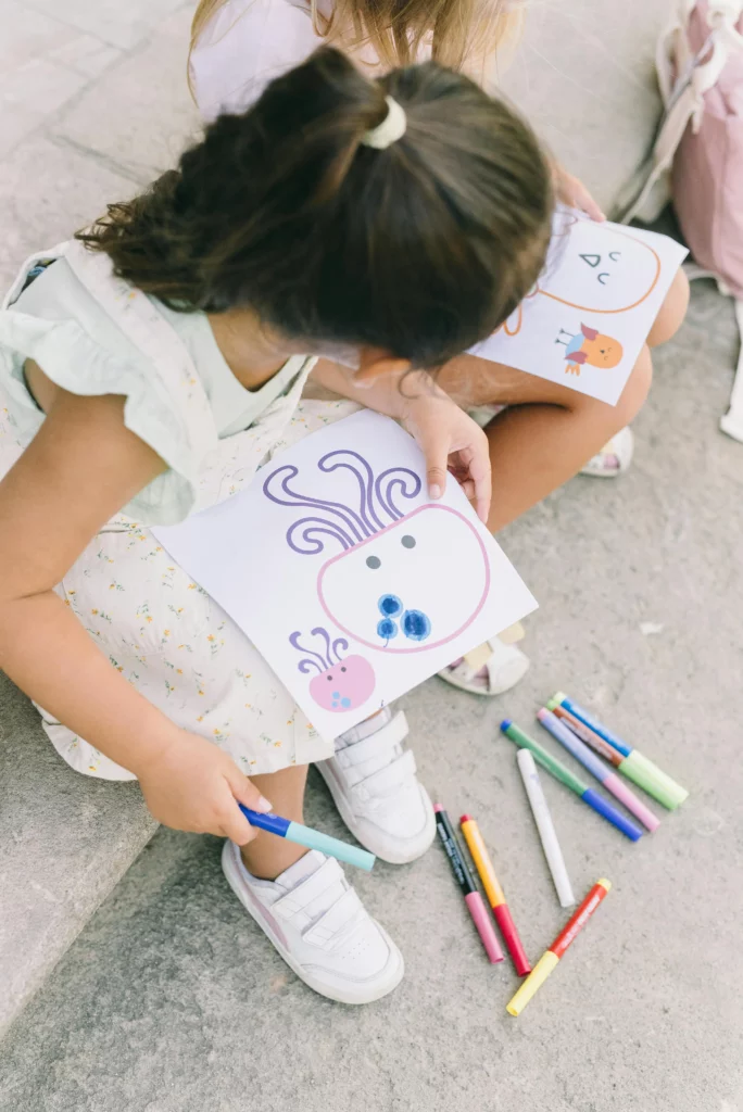 Two children sitting outdoors holding drawings of a cartoon octopus and bird with markers nearby.