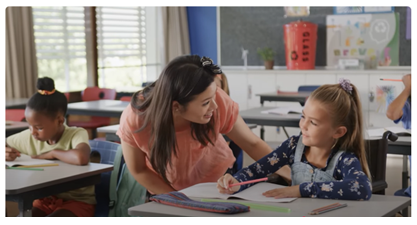 Teacher assisting a young student at her desk while another student works in the classroom.