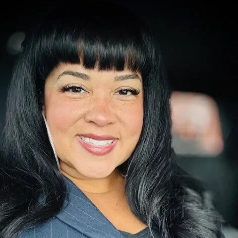 Woman with long black hair and bangs wearing a dark blazer and black top smiling at the camera