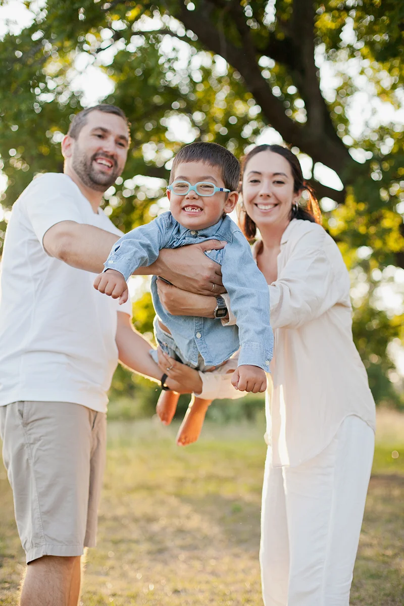 Family outdoors: a man and woman hold a smiling toddler with glasses between them.